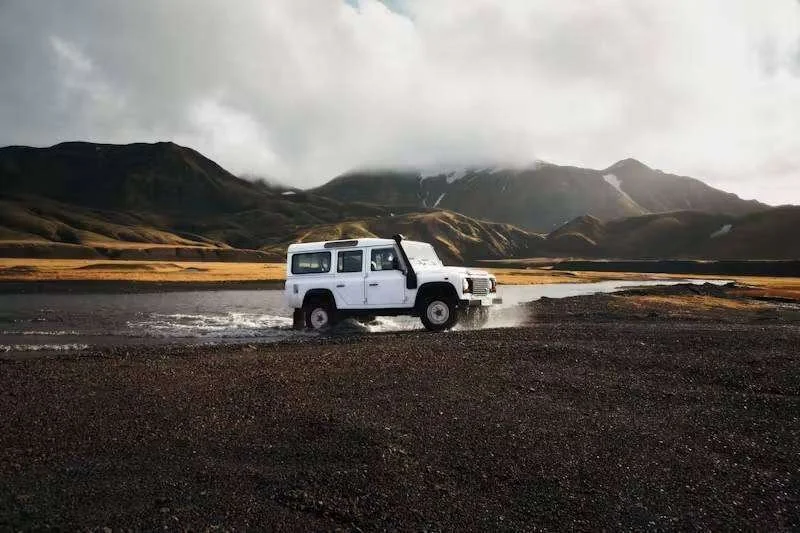Jipe 4x4 branco atravessa rio com respingos em paisagem montanhosa e céu nublado